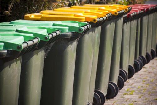 A row of large, green wheeled rubbish bins with black wheels and handles is aligned along a paved surface, with their bright yellow and red lids closed on top. The bins are made of durable plastic with a smooth finish and are positioned outdoors, possibly near a residential or commercial property in Chislehurst. The environment appears well-maintained, with a background of greenery and sunlight illuminating the scene. The composition emphasizes the uniformity and readiness of the waste containers, typical of rubbish removal services by Office Clearance Chislehurst operating within the local area, ensuring proper waste disposal and collection.