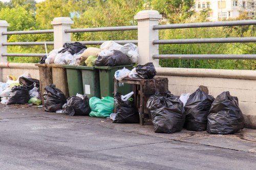 A collection of black, green, and white rubbish bags filled with waste, some loosely tied and others sealed, are piled against a concrete railing along a paved urban street. Behind the bags, there are three large wheelie bins in dark green and brown, with their lids closed, positioned on a sidewalk next to the railing. The scene appears to be part of an outdoor waste collection area in a residential or commercial area, with leafy trees and a building visible in the background. The bags and bins are situated on a dark asphalt surface, with the bags overflowing onto the pavement. The image reflects a typical rubbish removal situation in a suburban area, which local services like Office Clearance Chislehurst could assist with, particularly in managing excess waste and ensuring proper disposal in the Chislehurst area.