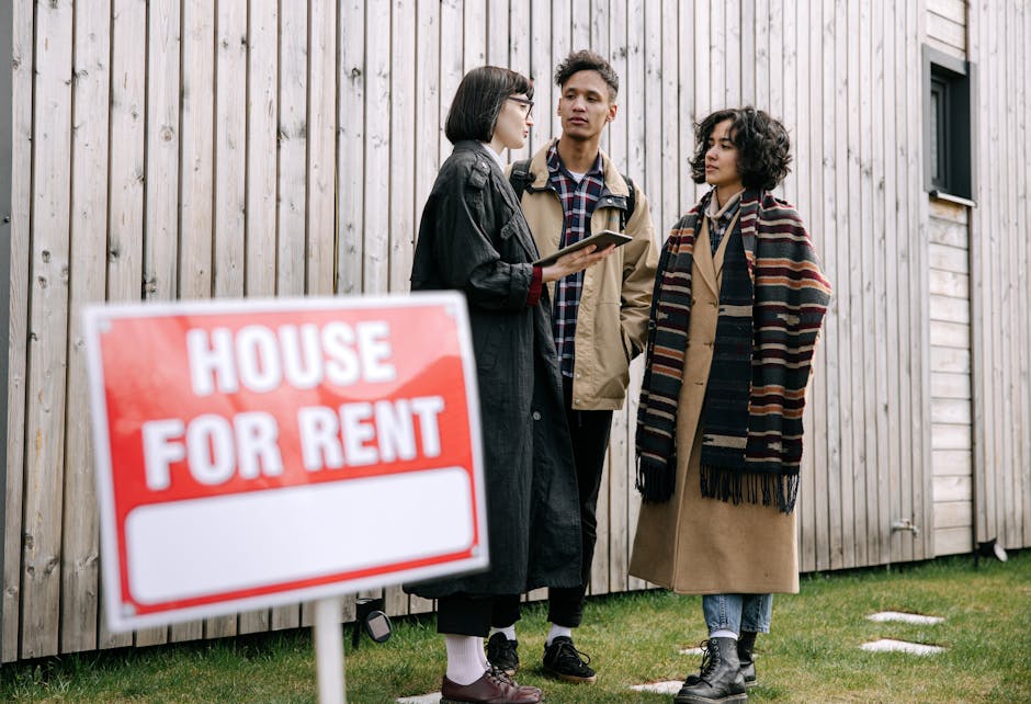 A red and white rectangular 'House For Rent' sign with bold capital letters is mounted on a wooden post positioned in front of a modern building exterior. The building features light-colored vertical