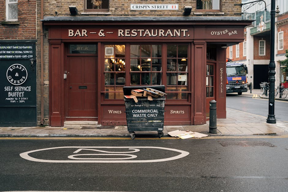 Four large wheeled rubbish bins are positioned in a row on a cobbled street in front of a peach-colored building with three barred window openings. The first bin, on the left, is dark green with a sli