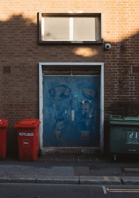 The image depicts a weathered, blue metal double door set into a red brick wall, with visible signs of rust and peeling paint. Above the door, there is a rectangular, horizontally oriented, frosted gl